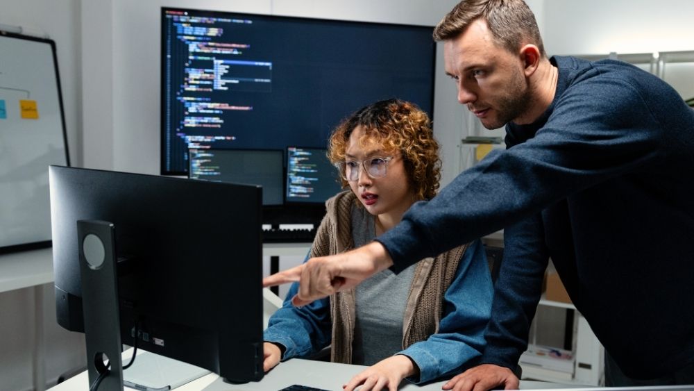 Two software developers reviewing code on a screen in a dark office, one pointing to a build pipeline display